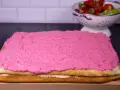 En tårtbotten i långpanna med rosa glasyr. I bakgrunden en skål med jordgubbar och kiwi.