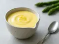 En skål med krämig hollandaisesås, garnerad med citronskal. Sparris och en sked syns i bakgrunden.