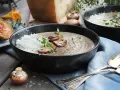 Creamy mushroom soup in a black bowl, topped with grated cheese, mushrooms, and herbs.