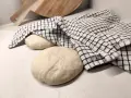 Two balls of pizza dough on a counter, one covered with a checkered cloth, with a wooden pizza peel in the background.