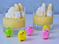 Två glas med gul pannacotta, toppade med vispgrädde och marshmallows som liknar påskkycklingar. Små, fluffiga påskkycklingar i olika färger står framför.
