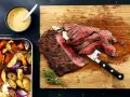 Sliced flank steak with roasted vegetables and a bowl of sauce.