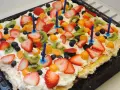A rectangular meringue cake topped with whipped cream, strawberries, kiwi, blueberries, and physalis, with blue birthday candles.
