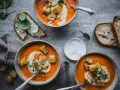 Three bowls of vibrant tomato soup topped with cream, croutons, and herbs, with bread and spinach on the side.
