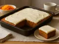 A rectangular carrot cake with white frosting in a baking pan, with a slice on a plate and a bowl of shredded carrots.