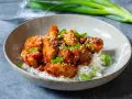 Krispig koreansk kyckling med sesamfrön och salladslök serveras på en bädd av vitt ris i en skål.