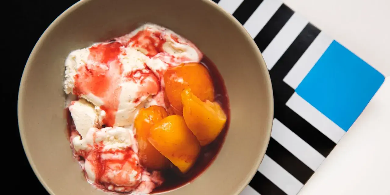 En skål med krämig vaniljglass, saftiga persikohalvor och en röd bärsås. En klassisk peach melba som ser utsökt ut.
