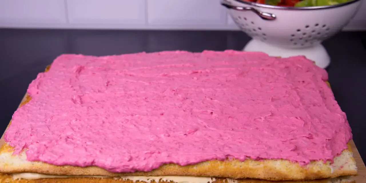 En tårtbotten i långpanna med rosa glasyr. I bakgrunden en skål med jordgubbar och kiwi.