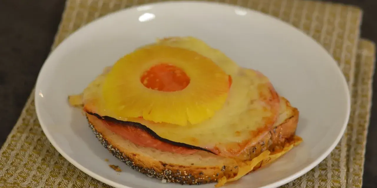 A toasted sandwich with melted cheese, ham, and a pineapple ring on top, served on a white plate.