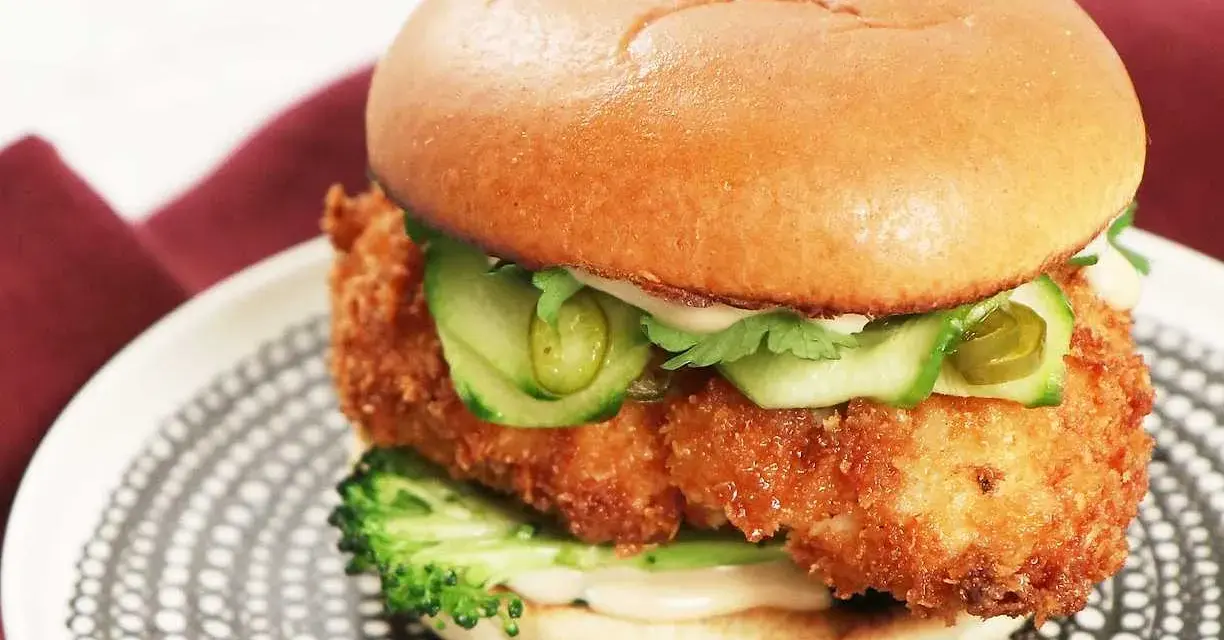 En saftig kyckling hamburgare med krispigt panerat kycklingfilé, gurka, jalapeños och en krämig sås på ett mjukt bröd. Serveras på en snygg tallrik.