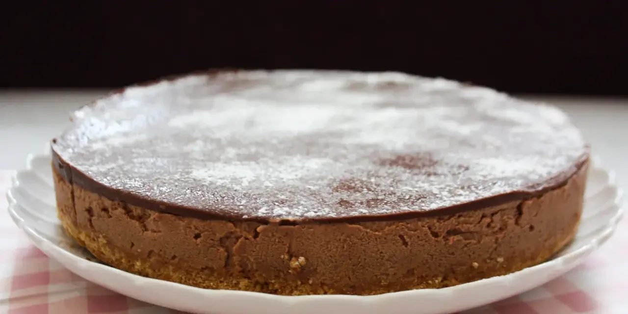 En krämig nutella cheesecake med chokladglasyr och pudersocker, serverad på en vit tallrik med en rosa rutig duk.
