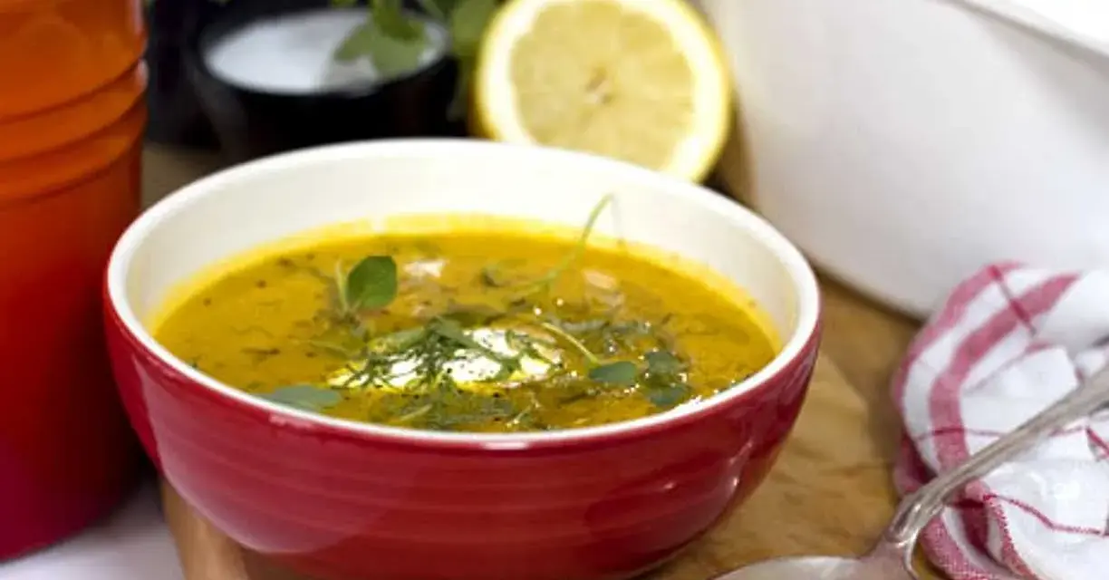 Varm morotssoppa med en klick creme fraiche, garnerad med örter. Serveras i en röd skål med en sked och en citronskiva i bakgrunden.