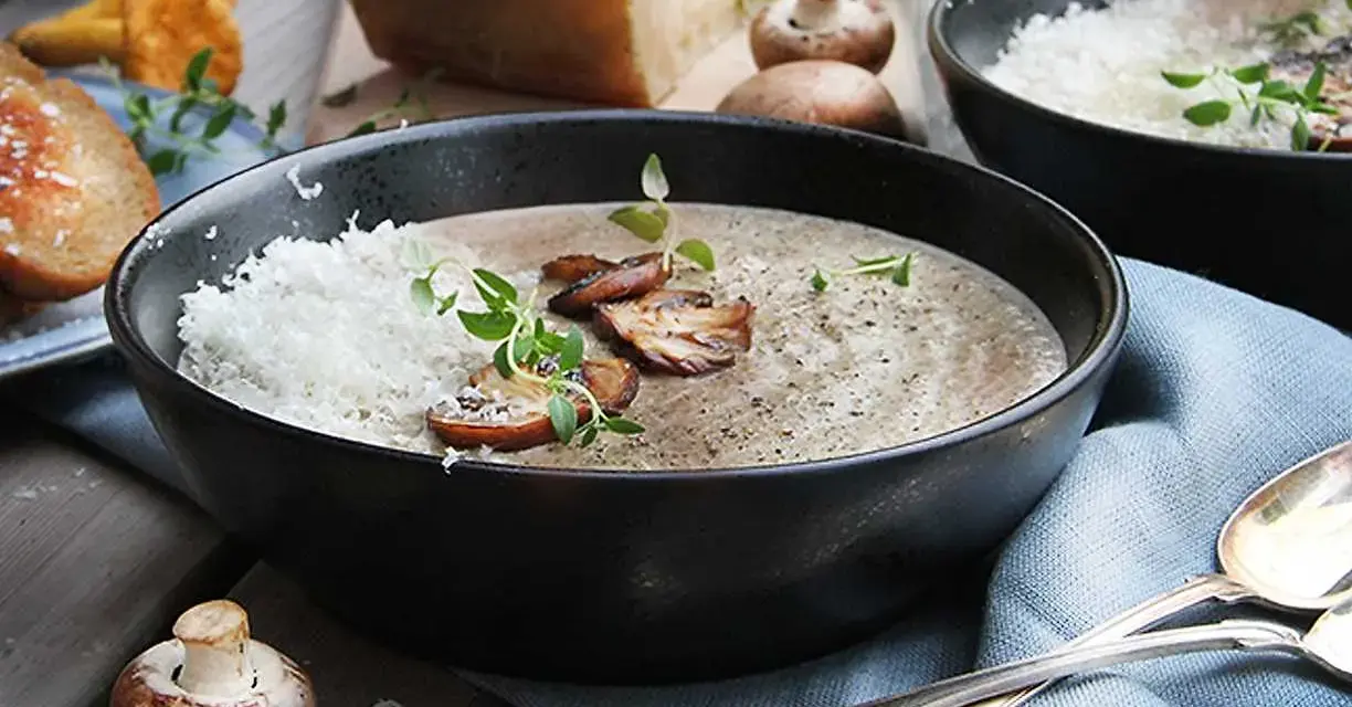 Creamy mushroom soup in a black bowl, topped with grated cheese, mushrooms, and herbs.