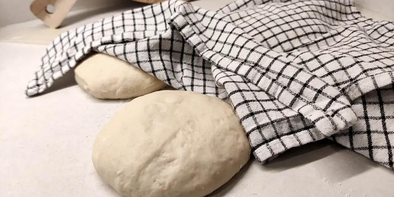 Two balls of pizza dough on a counter, one covered with a checkered cloth, with a wooden pizza peel in the background.