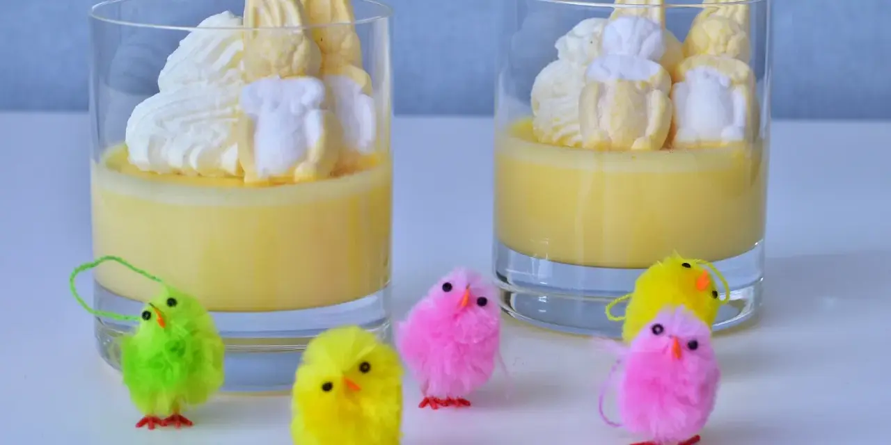 Två glas med gul pannacotta, toppade med vispgrädde och marshmallows som liknar påskkycklingar. Små, fluffiga påskkycklingar i olika färger står framför.