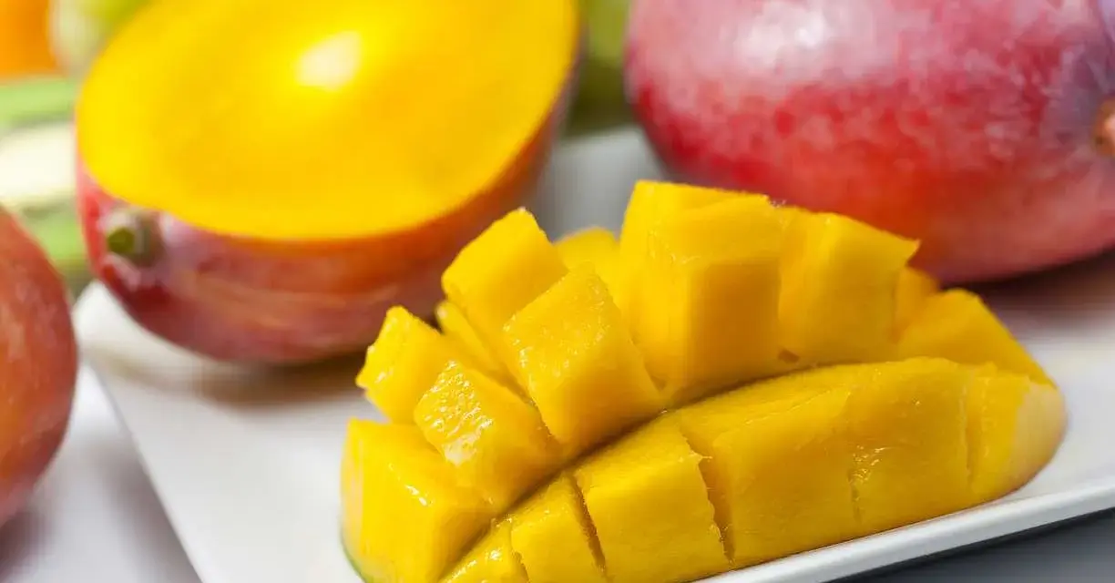 En hel och en halverad mango, skuren i bitar, ligger på en vit tallrik. Bakom syns druvor och en annan hel mango.