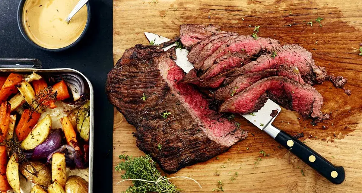 Sliced flank steak with roasted vegetables and a bowl of sauce.
