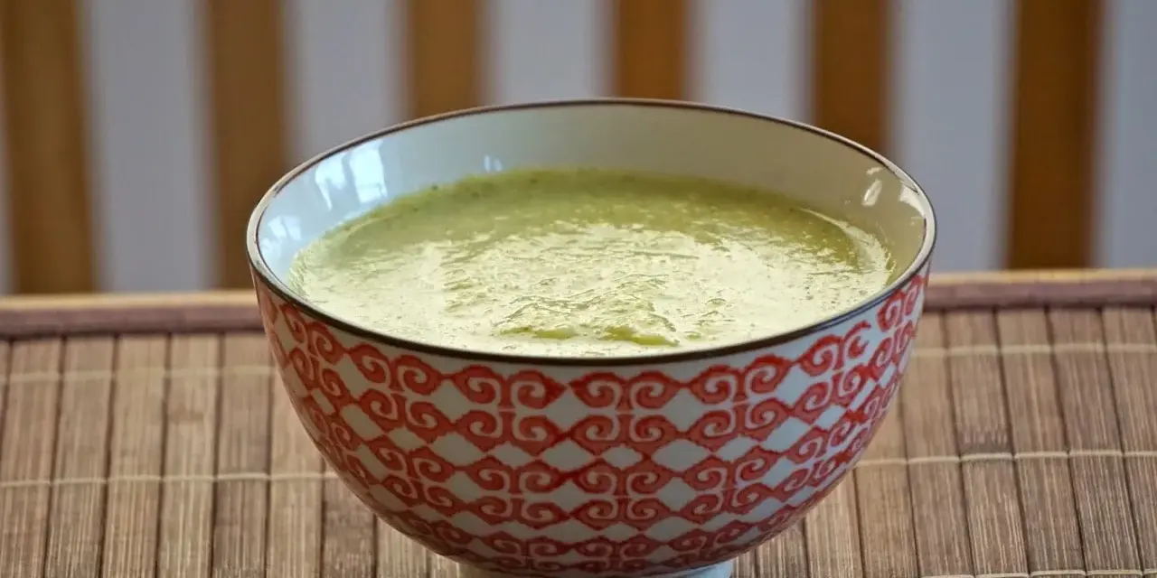 En skål med krämig blomkål och broccolisoppa, serverad på en bambumatta.