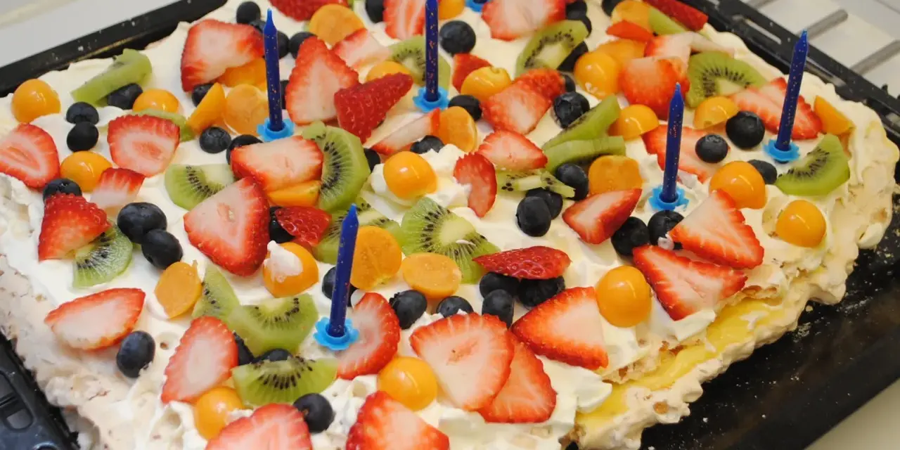 A rectangular meringue cake topped with whipped cream, strawberries, kiwi, blueberries, and physalis, with blue birthday candles.