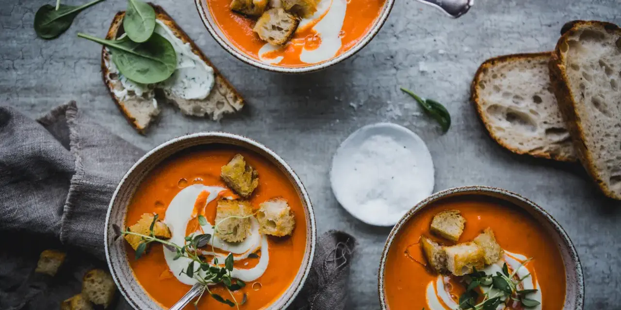 Three bowls of vibrant tomato soup topped with cream, croutons, and herbs, with bread and spinach on the side.
