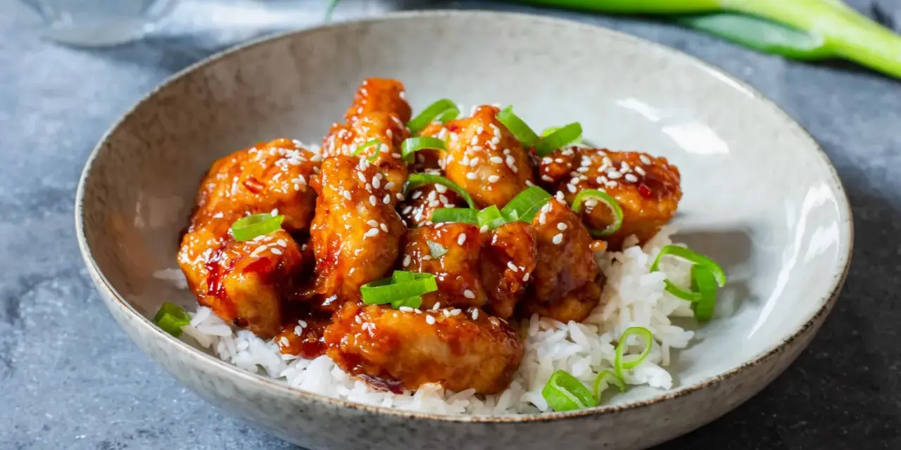 Krispig koreansk kyckling med sesamfrön och salladslök serveras på en bädd av vitt ris i en skål.