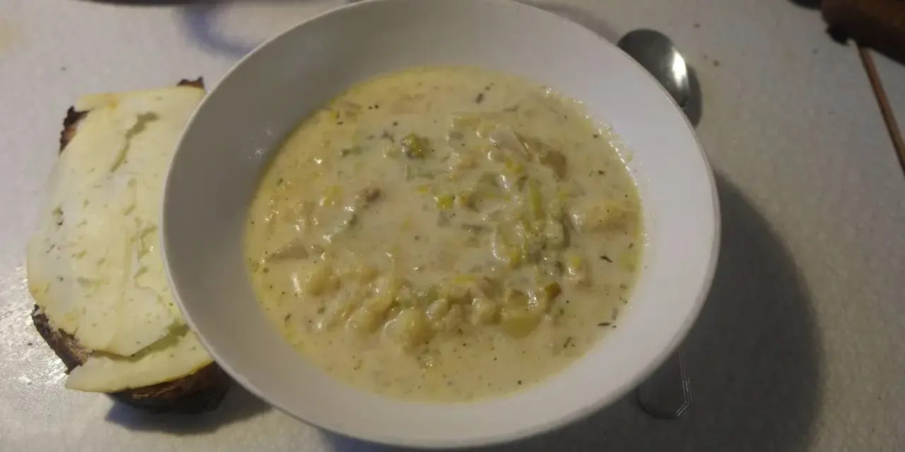 En krämig fänkålssoppa med potatis serveras i en vit skål, bredvid en bit rostat bröd med ost.