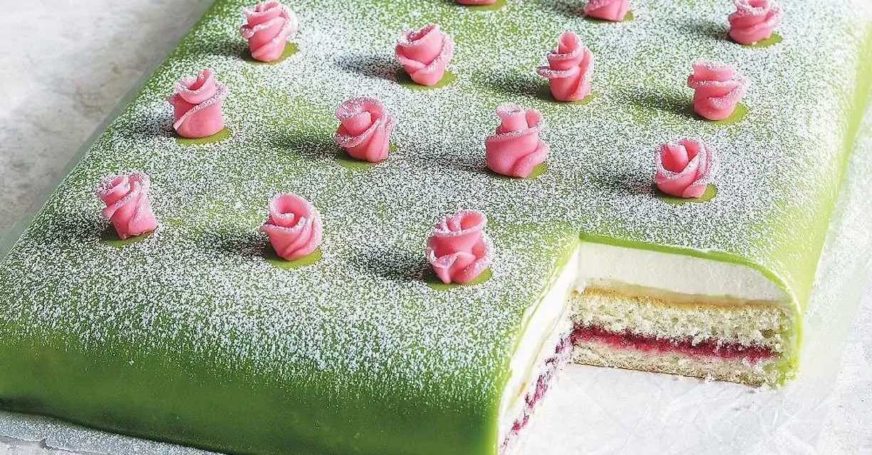 A rectangular green cake topped with powdered sugar and decorated with pink marzipan roses. A slice is cut out, revealing layers of cake, cream, and jam.