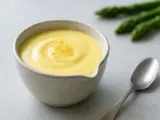 En skål med krämig hollandaisesås, garnerad med citronskal. Sparris och en sked syns i bakgrunden.