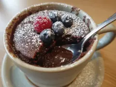Chokladig mug cake med hallon och blåbär, pudrad med florsocker. En sked dyker ner i den rinnande chokladen.