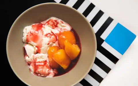 En skål med krämig vaniljglass, saftiga persikohalvor och en röd bärsås. En klassisk peach melba som ser utsökt ut.