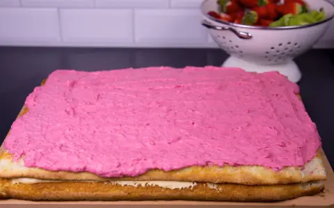 En tårtbotten i långpanna med rosa glasyr. I bakgrunden en skål med jordgubbar och kiwi.