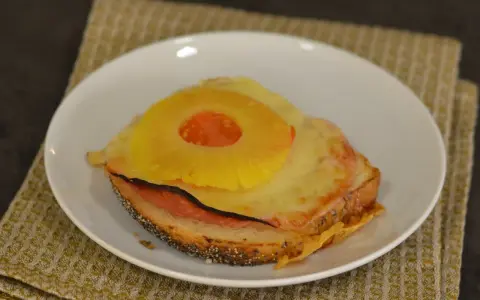 A toasted sandwich with melted cheese, ham, and a pineapple ring on top, served on a white plate.