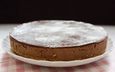 En krämig nutella cheesecake med chokladglasyr och pudersocker, serverad på en vit tallrik med en rosa rutig duk.