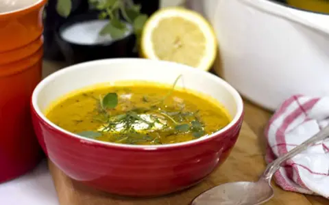 Varm morotssoppa med en klick creme fraiche, garnerad med örter. Serveras i en röd skål med en sked och en citronskiva i bakgrunden.