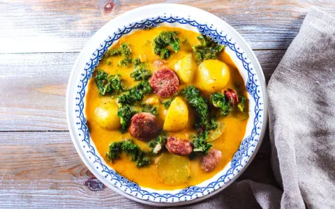 En mustig grönsakssoppa med potatis, grönkål och korvskivor i en gul buljong.