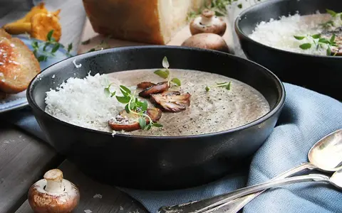 Creamy mushroom soup in a black bowl, topped with grated cheese, mushrooms, and herbs.