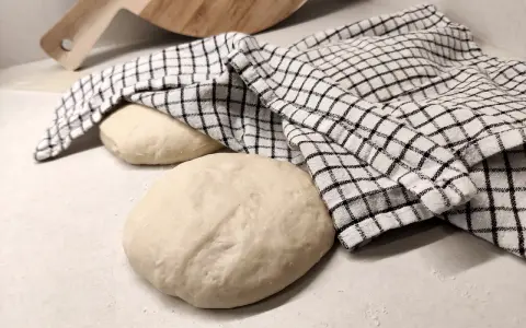 Two balls of pizza dough on a counter, one covered with a checkered cloth, with a wooden pizza peel in the background.