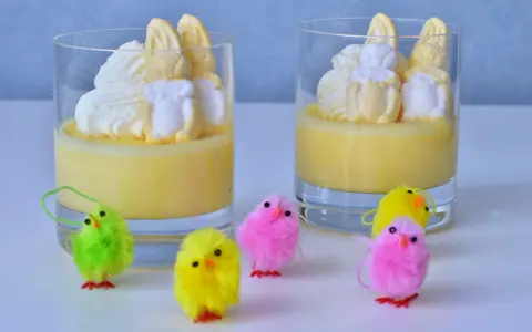 Två glas med gul pannacotta, toppade med vispgrädde och marshmallows som liknar påskkycklingar. Små, fluffiga påskkycklingar i olika färger står framför.