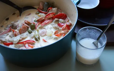 En krämig kreolsk gryta med korv, paprika, oliver och pickles i en blå gryta. En sked ligger i ett glas med mjölk.