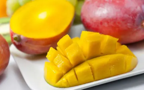 En hel och en halverad mango, skuren i bitar, ligger på en vit tallrik. Bakom syns druvor och en annan hel mango.
