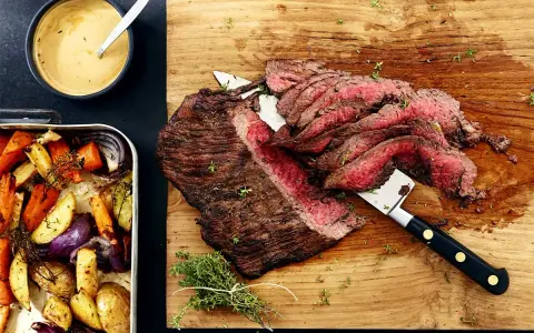 Sliced flank steak with roasted vegetables and a bowl of sauce.