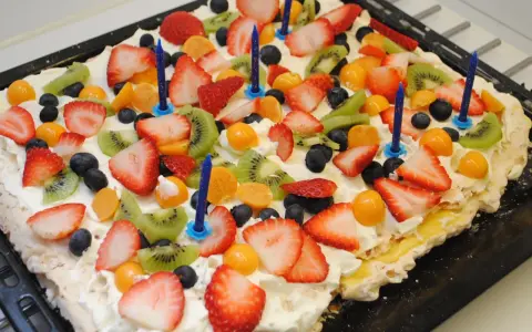 A rectangular meringue cake topped with whipped cream, strawberries, kiwi, blueberries, and physalis, with blue birthday candles.