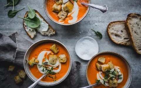 Three bowls of vibrant tomato soup topped with cream, croutons, and herbs, with bread and spinach on the side.