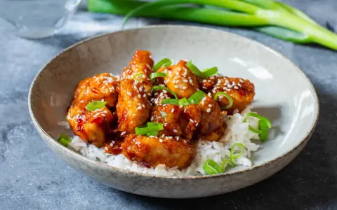 Krispig koreansk kyckling med sesamfrön och salladslök serveras på en bädd av vitt ris i en skål.