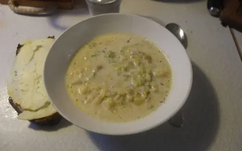 En krämig fänkålssoppa med potatis serveras i en vit skål, bredvid en bit rostat bröd med ost.