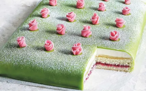 A rectangular green cake topped with powdered sugar and decorated with pink marzipan roses. A slice is cut out, revealing layers of cake, cream, and jam.