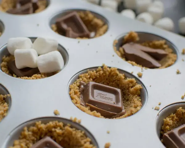 S'mores i ugn förbereds i en muffinsform med krossade kex, chokladbitar och marshmallows.