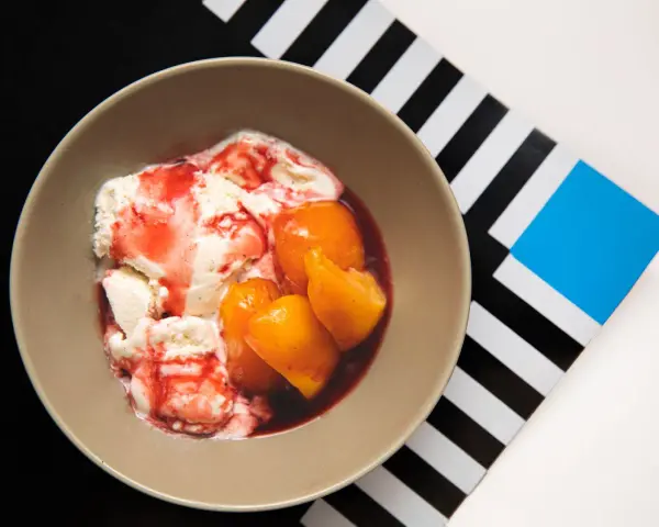 En skål med krämig vaniljglass, saftiga persikohalvor och en röd bärsås. En klassisk peach melba som ser utsökt ut.
