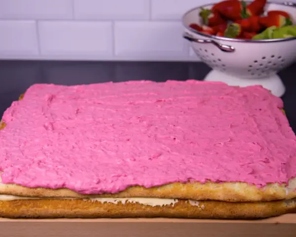 En tårtbotten i långpanna med rosa glasyr. I bakgrunden en skål med jordgubbar och kiwi.