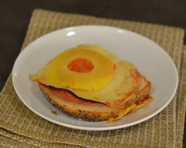 A toasted sandwich with melted cheese, ham, and a pineapple ring on top, served on a white plate.